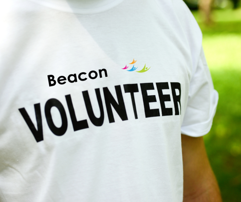 This image shows a person, whose head is cropped out the picture, wearing a white t-shirt with the words Beacon volunteer on it in white writing.