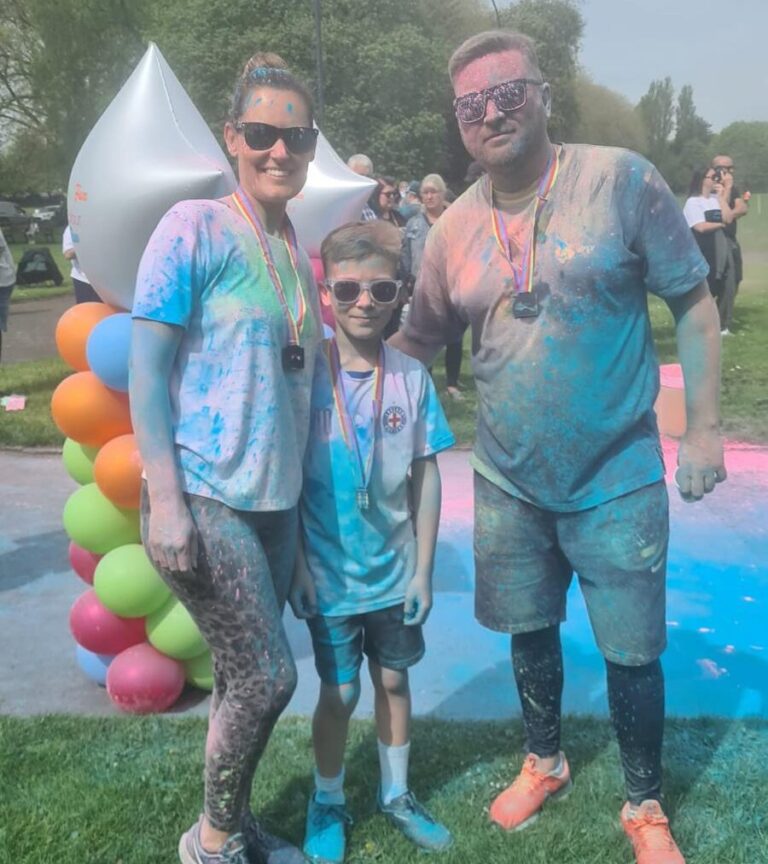 This shows three people, a man, woman and young child at a colour run in aid of Beacon. They are covered in brightly covered paint powder.